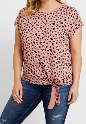 Kobieta w różowej koszulce na krótki rękaw w panterkę, zawiązanej na supeł w talii, z ciemnoniebieskimi podartymi jeansami.