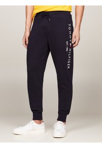 Marina blå sweatpants med dragsko i midjan, prydda med en vit "TOMMY HILFIGER"-logotyp längs sidan och ribbade muddar, tillsammans med vita sneakers.