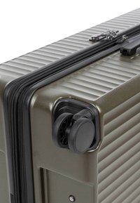 Coin d'une valise rigide vert olive avec des doubles roues pivotantes noires et une fermeture éclair noire le long du bord.