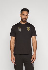 Camiseta negra con un logo bordado en dorado y el número "10" en la parte delantera. Mangas cortas y un diseño clásico de cuello redondo.