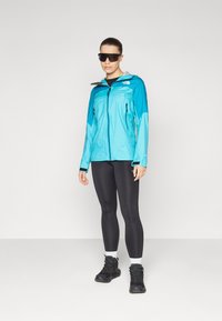 Letvægts blå regnjakke med grå detaljer, lynlåslommer og hætte, båret med sorte leggings og vandrestøvler.