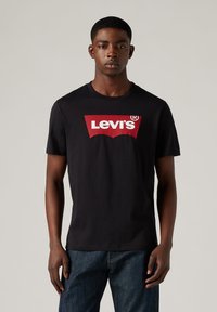Camiseta de algodón negra con un gran logo rojo de Levi's en el pecho. Mangas cortas, cuello redondo y un corte casual. Llevada con pantalones vaqueros azules.