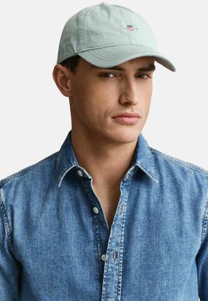 Uomo con cappellino grigio chiaro e camicia di jeans blu con bottoni, che guarda leggermente di lato con un'espressione neutra.