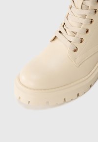 Bottine en cuir beige avec un bout arrondi, semelle plate en caoutchouc et lacets beiges ; présente des œillets dorés et une texture lisse.