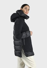 Sort pufferjakke med hætte, der har en blanding af glatte og quiltede strukturer, lynlåsede sider og et længere design bagtil for ekstra dækning.