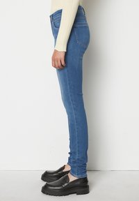 Jeans subțiri din denim albastru, cu talie înaltă, caracterizându-se printr-un design clasic cu cinci buzunare și un finisaj neted. Asortate cu pantofi negri, slip-on.