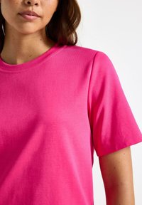 Nærbillede af en kvinde iført en ensfarvet lyserød t-shirt med korte ærmer mod en hvid baggrund.