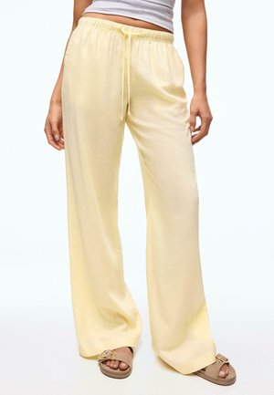 Person wearing loose, pale yellow drawstring pants and beige slide sandals with gold buckles, standing against a plain background.