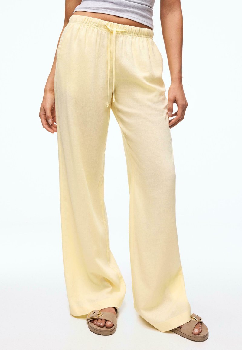 Personne portant un pantalon ample jaune pâle avec cordon et des sandales slides beiges avec boucles dorées, debout devant un fond uni.