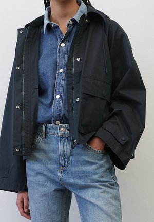 Personne portant une chemise en denim superposée et un jean avec une veste noire ample, une main dans la poche, sur un fond uni.