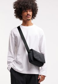 Sac en bandoulière noir en nylon avec une texture rembourrée et une fermeture par rabat, porté sur une chemise blanche à manches longues et associé à un pantalon noir.