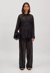 Schwarze durchsichtige Bluse mit Knöpfen, zwei Brusttaschen und langen Ärmeln, kombiniert mit passenden, locker sitzenden durchsichtigen Hosen und flachen Sandalen.