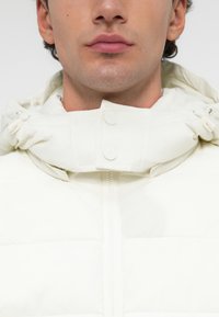 Λευκή φούτερ ζακέτα με ψηλό γιακά, διαθέτει μπροστινό φερμουάρ και κουμπιά με κλιπ. Ομαλή υφή με καπιτονέ τμήματα και λεπτομέρειες με κορδόνι.