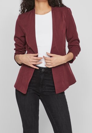 Vrouw die een aangesloten bordeauxrode blazer draagt over een wit topje en een zwarte jeans met hoge taille, en de blazer met beide handen rechtzet.