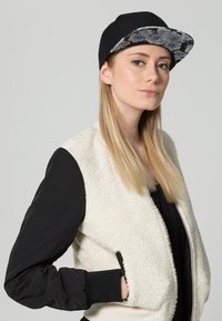 Weißer Fleece-Bomberjacke mit schwarzen Ärmeln, kombiniert mit einer schwarzen Kappe mit einem blumenmusternden Schirm. Model von der Seite blickend.