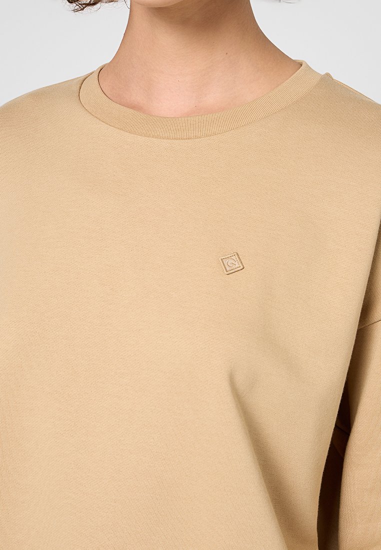 Sudadera de algodón beige con escote amplio, hombros caídos y un pequeño logo bordado en el pecho. Textura suave y lisa.