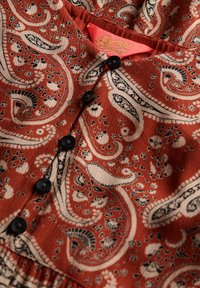 Tkanina w kolorze rdzy z wzorem paisley w kremowym i czarnym; ma czarne guziki i różową metkę ze złotym tekstem.