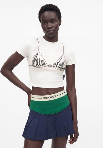 Γυναίκα που φοράει λευκό crop top με τύπωμα σουτιέν "Palm Angels", πράσινη ζώνη μέσης με κείμενο "Palm Angels" και σκούρο μπλε πλισέ φούστα, στέκεται με το χέρι στη μέση.