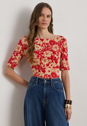 Γυναίκα με μακριά κυματιστά μαλλιά που φοράει κόκκινη και μπεζ φλοράλ μπλούζα, μπλε τζιν παντελόνι με φαρδιά μπατζάκια και μεγάλα χρυσά και καμηλοπαρδάλεα βραχιόλια σε έναν καρπό.