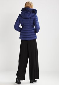Námořnicky modrá péřová bunda s límcem z umělé kožešiny a horizontálním prošíváním. Kombinována s širokými černými kalhotami a kotníkovými botami.