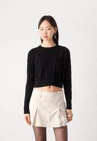 Svart kortärmad tröja med texturerad ribbning och kabelstickad design, tillsammans med en ljusbeige minikjol. Modellen bär svarta strumpbyxor.