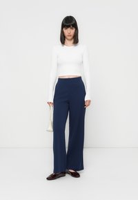 Witte lange mouwen cropped top, marineblauwe wijde broek en bordeauxrode schoenen. Model houdt een kleine witte handtas vast tegen een eenvoudige achtergrond.