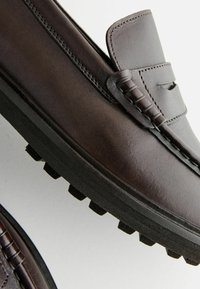 Mocasines de cuero marrón oscuro con un acento de costura y detalle elevado, que presentan una suela de goma texturizada para agarre y durabilidad.