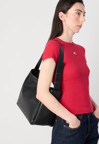 Une femme aux longs cheveux noirs porte une chemise rouge et tient un sac à main noir, se tenant devant un fond uni.