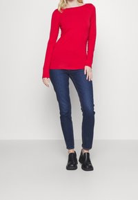 Camicia rossa a maniche lunghe con scollatura a barca, abbinata a jeans slim-fit blu e scarpe slip-on nere, ambientata su uno sfondo neutro.