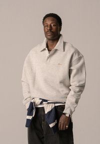 Grauer Pullover mit großem Kragen, gesticktem Logo und gerippten Bündchen. Blau-weißer Strickpullover um die Taille gebunden. Dunkle Jeans.