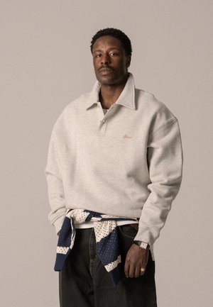Grå hettegenser med stor krage, brodert logo og ribbede mansjetter. Blå og hvit strikket genser knyttet rundt livet. Mørke jeans.