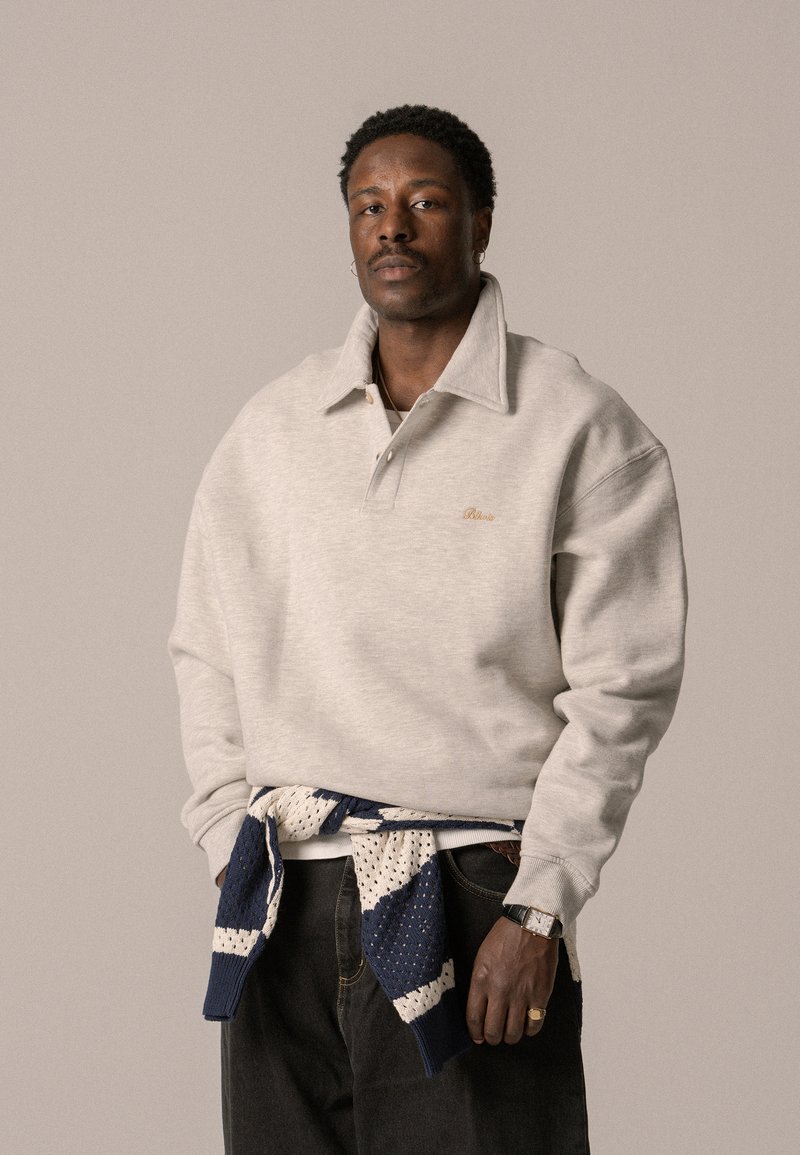 Grauer Pullover mit großem Kragen, gesticktem Logo und gerippten Bündchen. Blau-weißer Strickpullover um die Taille gebunden. Dunkle Jeans.