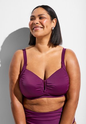 Žena srednje boje kože i ravne tamne kose koja se smiješi, nosi ljubičasti gornji dio bikinija i odgovarajuće donje dijelove, stoji na bijeloj pozadini.