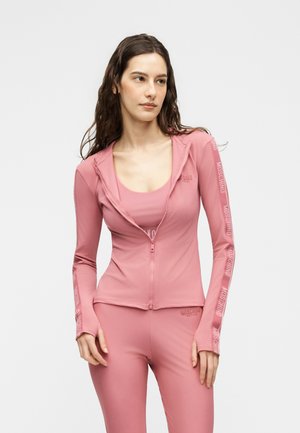 Žena s dlhými hnedými vlasmi, ktorá má na sebe priliehavú ružovú bundu na zips a zodpovedajúce legíny s textom loga na rukávoch a v páse.