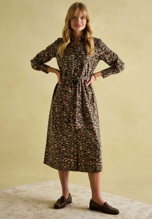 Langes, grün gemustertes Kleid mit Knopfleiste, Taillengürtel und langen Ärmeln. Kombiniert mit braunen Loafern auf einer strukturierten Oberfläche.