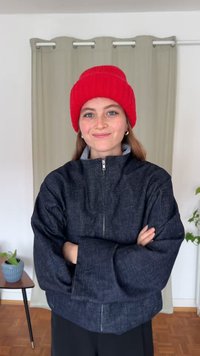 Tamsios džinsinės striukė su aukštu apykakliu ir sidabro spalvos užtrauktuku, derinama su ryškiai raudona megzta kepure. Fone matyti šviesios užuolaidos.