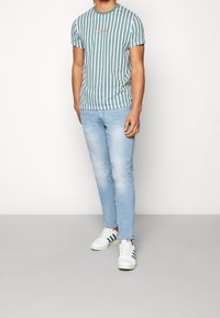 T-shirt w paski w kolorze turkusowym z białymi pionowymi paskami i graficznym napisem, w zestawieniu z jasnoniebieskimi dżinsami o slim fit i białymi trampkami z zielonymi akcentami.