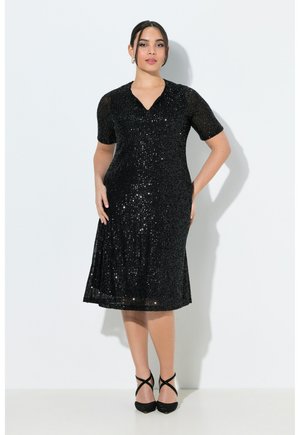 Kvinde stående iført en knælængde sort palietkjole med korte ærmer og sorte strop-sandaler mod en hvid baggrund.