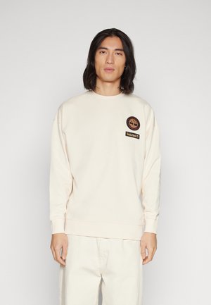 Sudadera color crema con puños y dobladillo acanalados, que presenta un parche de logo redondo y una etiqueta rectangular en el pecho. Textura suave.