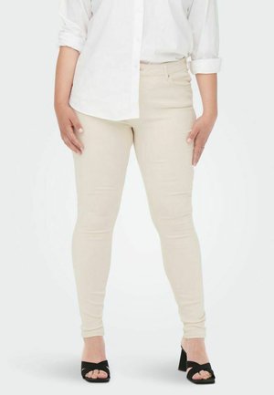 Frau trägt beige Skinny Jeans, weißes Hemd mit hochgekrempelten Ärmeln und schwarze offene Sandaletten mit Absatz, steht mit entspannten Händen.