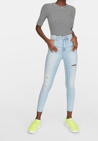 Persona vestida con una camiseta a rayas blanco y negro, jeans ajustados de mezclilla rotos color azul claro y zapatillas de deporte amarillas fluorescentes, de pie con las manos en las caderas.