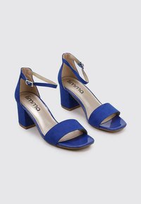 Sandalias de tacón alto azules con parte superior de ante, diseño destalonado, tira en el tobillo y tacón cuadrado brillante. Forro y plantilla en tonos neutros.