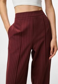 Femme portant un pantalon bordeaux à rayures verticales taille haute avec des poches latérales, associé à un haut court blanc, les mains reposant sur les côtés.