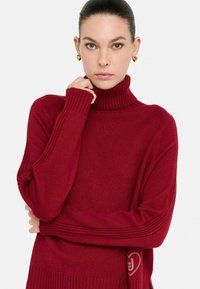 Rød turtleneck sweater med ribbet tekstur og lange ærmer. Har et hjerte-design og stripet detaljering nær kanten.