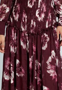 Vestido de flores burdeos con diseño plisado y cintura con volantes. La tela presenta un brillo sutil con grandes patrones de flores rosas claras.