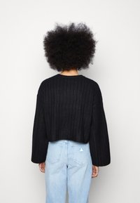 Czarny, skrócony sweter z prążkowanego dzianiny z szerokimi rękawami, z teksturowanym wzorem. Noszony na jasnoniebieskich jeansach o wysokim stanie.