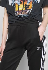 Svarta adidas träningsbyxor med vita sidostråk, elastisk midja och fickor med dragkedja. Tyget har en slät textur och en strukturerad passform.