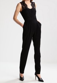 Svart jumpsuit med spetstopp, ärmlös design, skräddarsydd passform och avsmalnande ben med sidofickor. Matchas med svarta klackar.