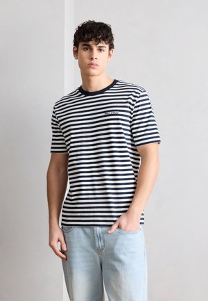 Tânăr cu păr creț poartă un tricou cu mânecă scurtă alb-negru în dungi și blugi albaștri deschis, stând în fața unui perete simplu, deschis la culoare.