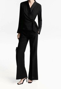 Blouse noire portefeuille à manches longues associée à un pantalon noir taille haute. Présente une texture lisse et une silhouette élégante. Talons noirs à brides.